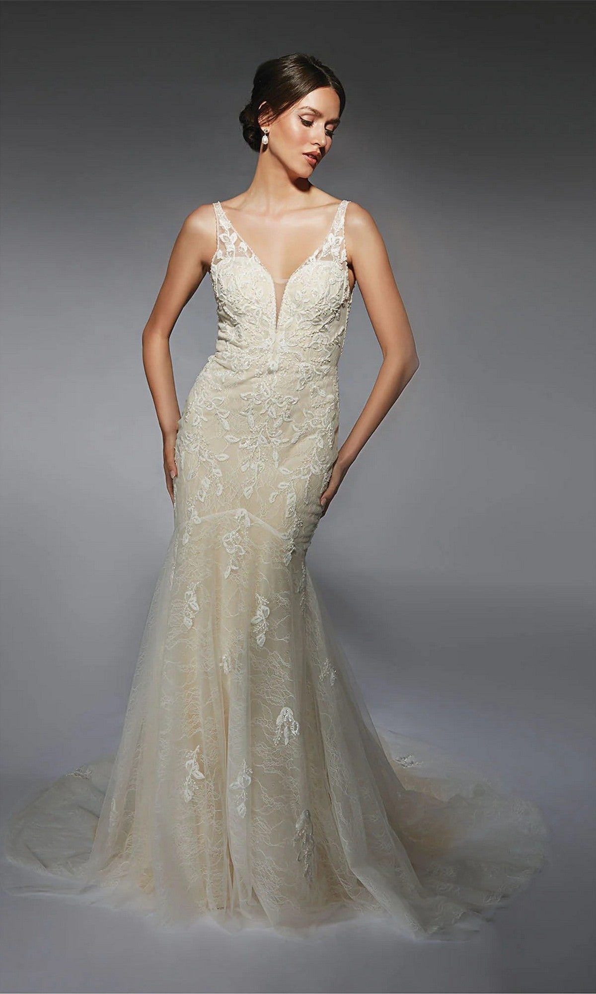 A woman poses against a gray background in the Alyce 7119 long white dress in ivory/champagne, a mermaid bridal gown with beaded lace, sheer straps, a deep V neckline, and delicate floral embroidery.